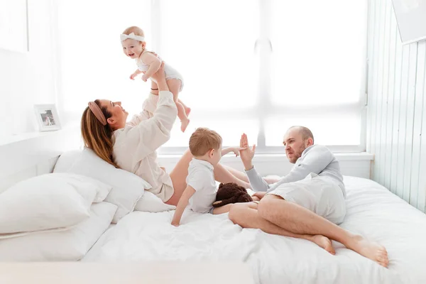 A young happy family with young children lying around, hugging, laughing on the bed in home clothes in a bright cozy house, mom, dad, son and daughter baby