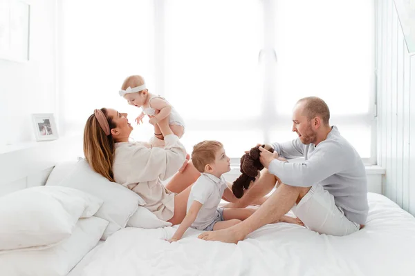 A young happy family with young children lying around, hugging, laughing on the bed in home clothes in a bright cozy house, mom, dad, son and daughter baby