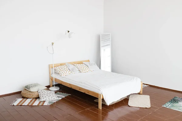 Yatak odasının içi. Geniş, parlak, beyaz duvarları ve ahşap zemini olan, küçük halıları, hasır koltuğu ve zemin lambası olan, ferah bir yatak odası.