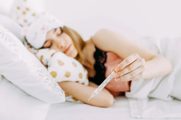 Pembe pijamalı ve kafasında battaniyeye sarılı uyku maskesi olan genç hasta bir kadın evde, yatak odasında, hasta olduğu için ilaç alıyor ve çay içiyor.