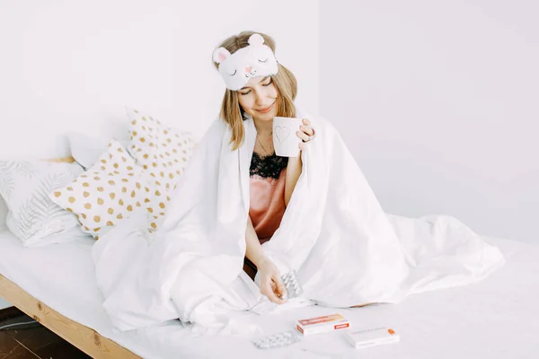 Pembe pijamalı ve kafasında battaniyeye sarılı uyku maskesi olan genç hasta bir kadın evde, yatak odasında, hasta olduğu için ilaç alıyor ve çay içiyor.