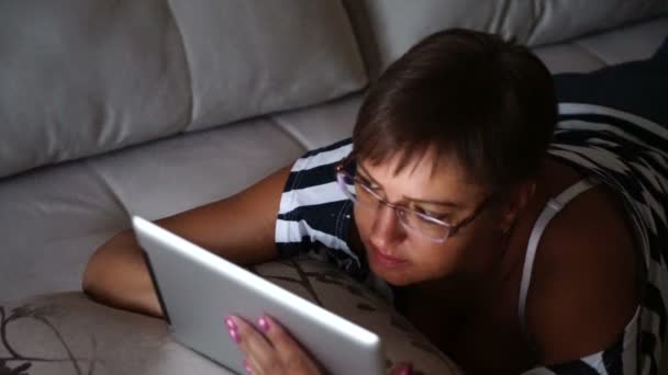 年轻女人浏览躺在沙发上在家里的平板电脑上的照片 — 图库视频影像