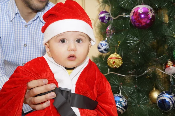Sehr niedlicher kaukasischer kleiner Junge im Weihnachtsmannkostüm mit Weihnachtsbaum und Dekoration im Hintergrund. Junge schaut in die Kamera. Weihnachtskonzept — Stockfoto
