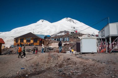 Karabashi ana kampı, Kafkasya, Rusya - Ağustos 2019: Güneşli bir günde Elbrus volkanının kayalık yamaçlarındaki ahşap ev ve diğer binaların yakınındaki insanlar.