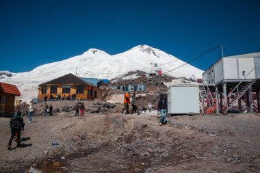 Karabashi ana kampı, Kafkasya, Rusya - Ağustos 2019: Güneşli bir günde Elbrus volkanının kayalık yamaçlarındaki ahşap ev ve diğer binaların yakınındaki insanlar.