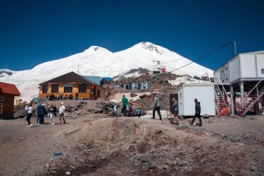 Karabashi ana kampı, Kafkasya, Rusya - Ağustos 2019: Güneşli bir günde Elbrus volkanının kayalık yamaçlarındaki ahşap ev ve diğer binaların yakınındaki insanlar.