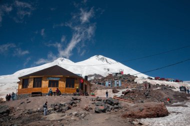 Karabashi ana kampı, Kafkasya, Rusya - Ağustos 2019: Güneşli bir günde Elbrus volkanının kayalık yamaçlarındaki ahşap ev ve diğer binaların yakınındaki insanlar.