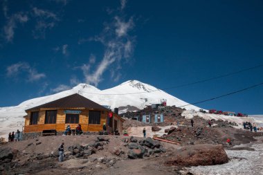 Karabashi ana kampı, Kafkasya, Rusya - Ağustos 2019: Güneşli bir günde Elbrus volkanının kayalık yamaçlarındaki ahşap ev ve diğer binaların yakınındaki insanlar.