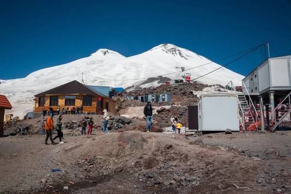 Karabashi ana kampı, Kafkasya, Rusya - Ağustos 2019: Güneşli bir günde Elbrus volkanının kayalık yamaçlarındaki ahşap ev ve diğer binaların yakınındaki insanlar.
