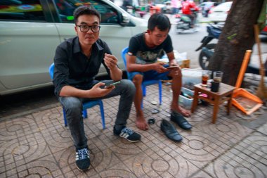 Ho Chi Minh şehri, Vietnam - Aralık 2018: Yoldaki küçük mavi sandalyelerde oturan ve cep telefonlarını izleyen iki Vietnamlı adam.