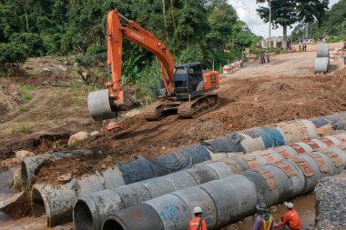 Chiangrai / Tayland - 20 Ağustos 20018, sel sonrası yol rehabilitasyonu. Bir turuncu kazıcı tesis için beton bir boru taşıyor, üç işçi izliyor, Chiangrai - Chiangmai yolu 118