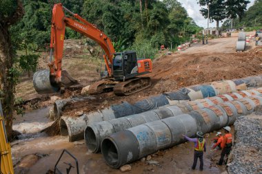 Chiangrai / Tayland - 20 Ağustos 20018, sel sonrası yol rehabilitasyonu. Bir turuncu kazıcı tesis için beton bir boru taşıyor, üç işçi izliyor, Chiangrai - Chiangmai yolu 118