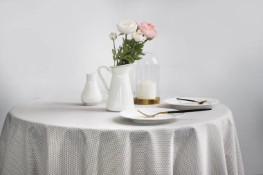 Çiçekli yuvarlak masa, hafif masa örtüsü ve hafif arka planda tabaklar..