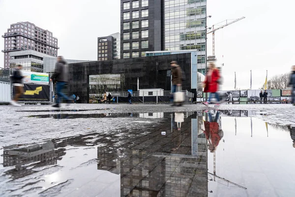People walking across a square in an urban setting reflected in a puddle of water
