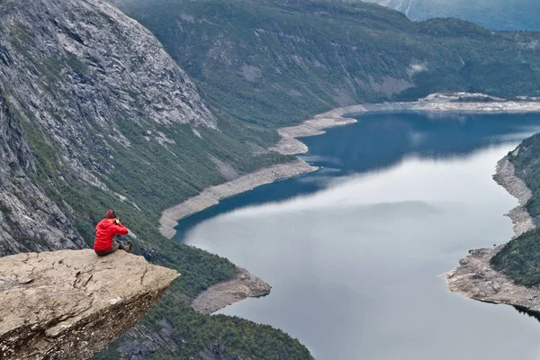 Homem com câmera sentada na rocha Trolltunga (Rocha da Língua do Troll) e faz a foto com a paisagem montesa norueguesa — Fotografia de Stock