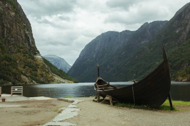 Nehrin arka planında eski Norveç Viking teknesi, dağlar ve mavi gökyüzü.