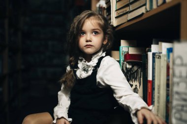 Okul üniformalı bir kız kitaplarla, okulla, librar arasında bir taburede oturuyor.