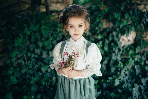 Yeşil-beyaz elbiseli kız elinde çiçekle eski bir taş duvarın ve ağacın arka planında.