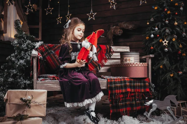a girl in Christmas costumes holds a nutcracker against the background of a bench, Christmas tree and a withered wall with stars