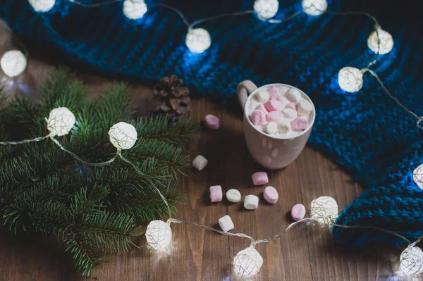 Cozy winter home. Cup of cocoa with marshmallows, blue knitted sweater, sprig of Christmas tree, garland, snowflakes on a white wooden table. Atmosphere of warm winter evening.