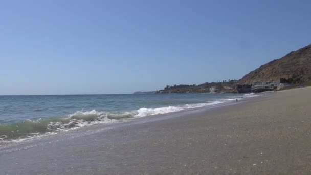 Fale pacyficznego oceanu biją na brzegu w Malibu Beach Los Angeles California. Kamera przesuwa się nad piaskiem. Stabilny strzał. Ciepły słoneczny letni dzień na plaży w Malibu. — Wideo stockowe