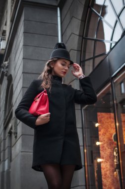 Alışveriş merkezinin cam duvar için seyir siyah şapka ve deri ceket, giyen genç güzel kız Kırmızı çantalı