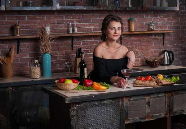 Beautiful woman with red wine glass sitting in kitchen  vegetables on table. — Stock Photo, Image