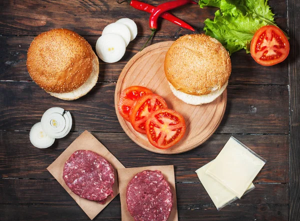 Homemade hamburger ingredients. Raw minced beef, fresh bun, slice of cheese, tomato, onion rings, lettuce on wood background