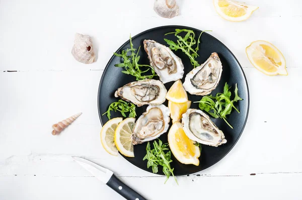 Ostriche con limone su un piatto nero su un tavolo di legno bianco — Foto Stock