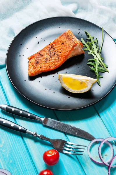 Один кусок запеченного лосося с лимоном на черной тарелке. Голубой фон — стоковое фото