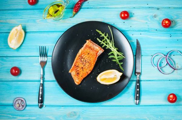 Un morceau de saumon cuit au four avec du citron sur une assiette noire. Fond en bois bleu — Photo