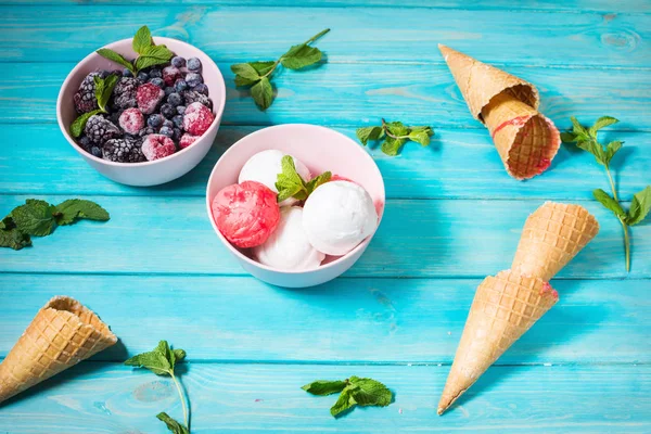 Ice cream in bowl, berries and waffle cones on blue wooden table. Summer