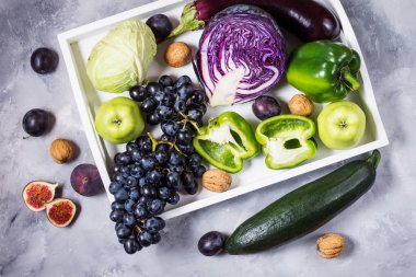 Taze organik çiğ yeşil ve mor renkli sebze ve meyve içinde karanlık taş zemin üzerine beyaz tepsi