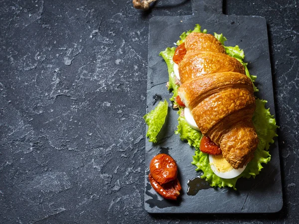 Sandwich de croissant con atún, huevo duro, ensalada y tomates secados al sol sobre mesa de piedra — Foto de Stock
