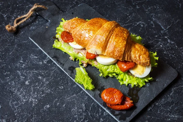 Sandwich de croissant con atún, huevo duro, ensalada y tomates secados al sol sobre mesa de piedra — Foto de Stock