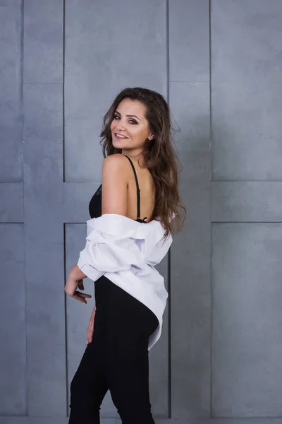 Portrait of a young smiling woman on a gray wall background. A beautiful girl in white shirt and black lingerie stands near a concrete wall — Stock Photo, Image