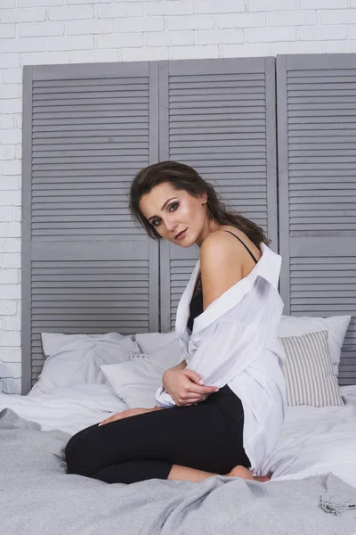Uzun saçlı bir kız siyah kot ve yatağın üstüne oturan beyaz gömlek giymiş. Şık casual kıyafet