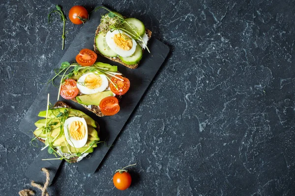 Tasty sandwiches with egg, avocado and vegetables on stone black background. Copy