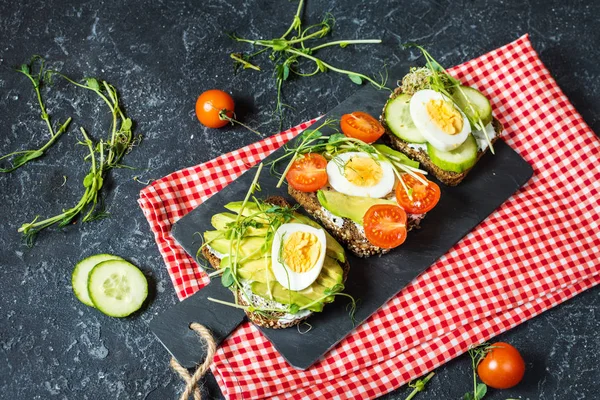 Tasty sandwiches with egg, avocado and vegetables on stone black background.