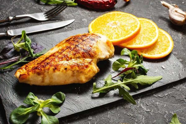 Grilled chicken fillets with orange on slate plate. Black stone background.