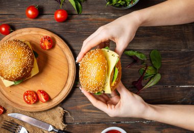 Kadın eli, ahşap arka planda tavuk, sahanda yumurta ve peynirli bir burger tutuyor. Üst görünüm