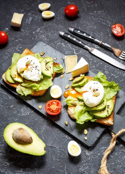 Healthy sandwich - poached eggs and avocado on toast with tomatoes on a black stone background.