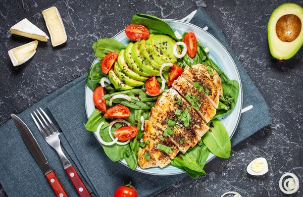 Grilled chicken breast and avocado salad with spinach and cherry tomatoes on black stone background. Healthy food, ketogenic diet, lunch concept. Top view