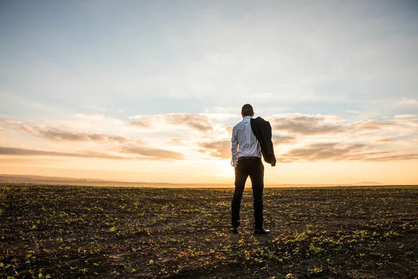 Empresário em terno elegante com seu casaco pendurado no campo durante o pôr do sol — Fotografia de Stock