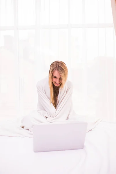 Привлекательная молодая женщина лежит на кровати под одеялом и с помощью ноутбука — стоковое фото