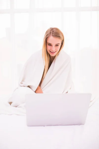 Привлекательная молодая женщина лежит на кровати под одеялом и с помощью ноутбука — стоковое фото