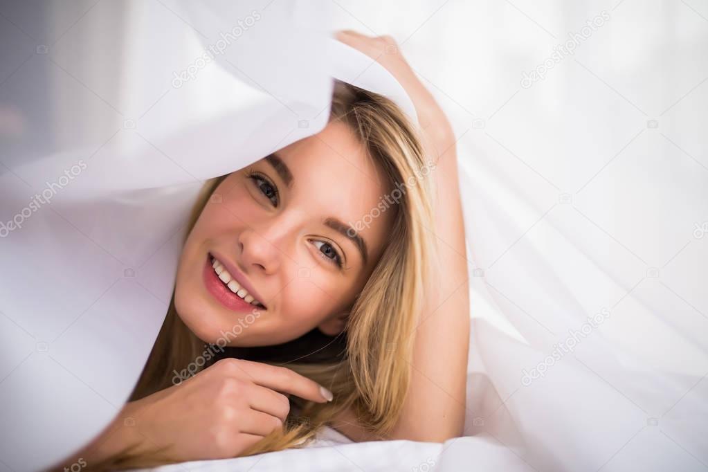 Smiling woman under a duvet in her bedroom. Closeup portrait of a beautiful young woman with blonde hair and under the blanket. happy good morning