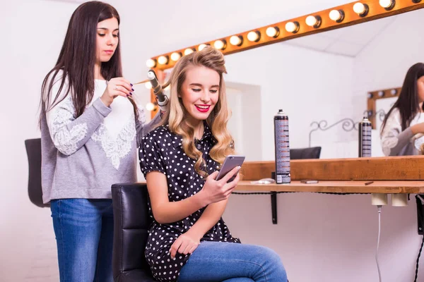 Female hairdresser applying hair straightener for long hair of smiling young woman using smartphone in dressing room