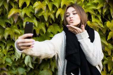 Genç kız gönder öpücük gülümseme ve selfie yeşil yaprakları arka plan üzerinde telefon almak