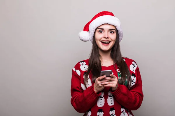 Menina modelo desgaste santa chapéu e Natal suéter texto sms no telefone no fundo cinza — Fotografia de Stock